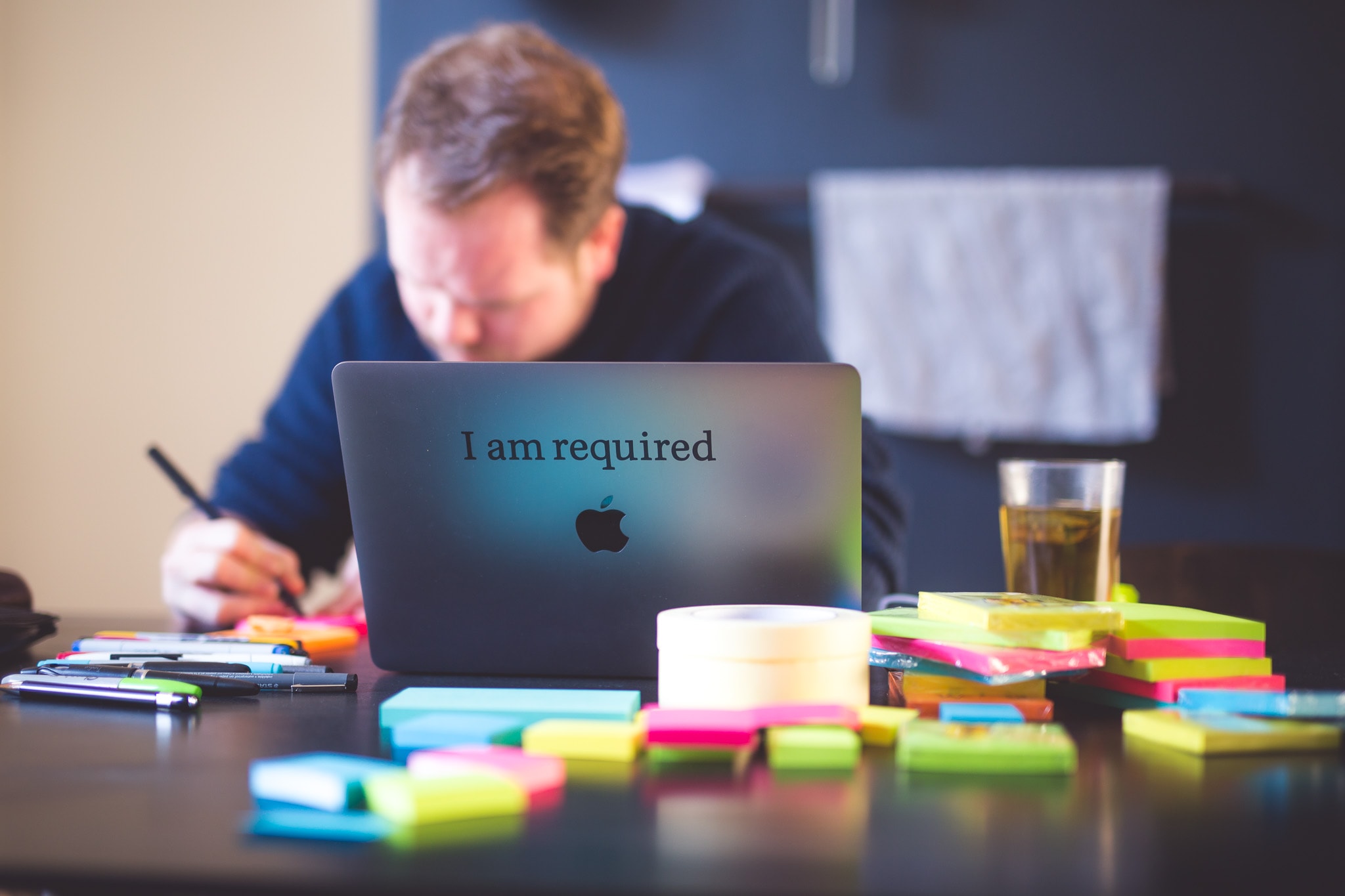 A laptop with the sticker "I am required". In the foreground you can see colorful Post-It notes, in the background Velthy writes something on paper