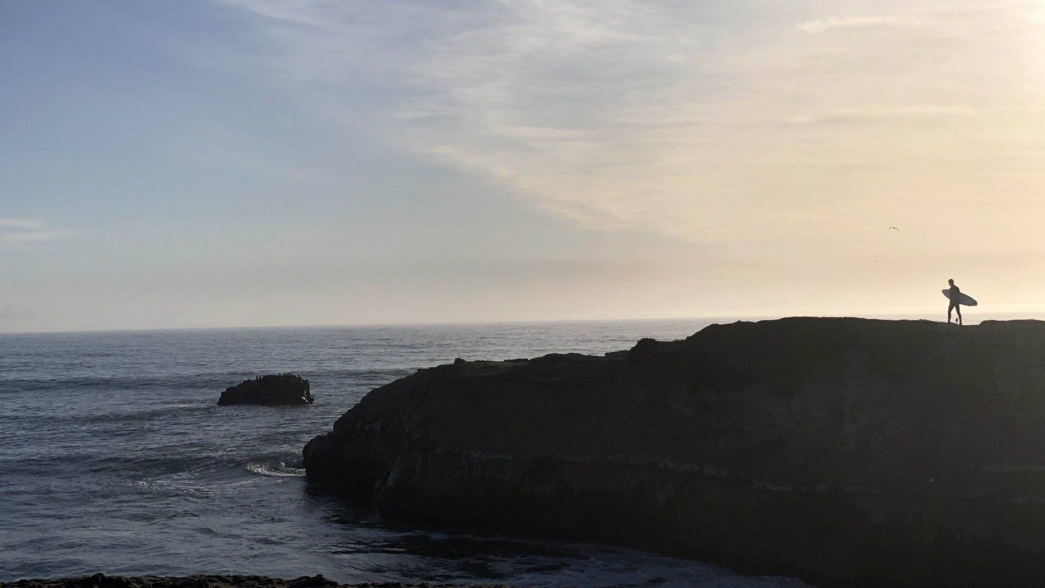 A cliff, right by the sea. A person with a surfboard can be seen in the background.
