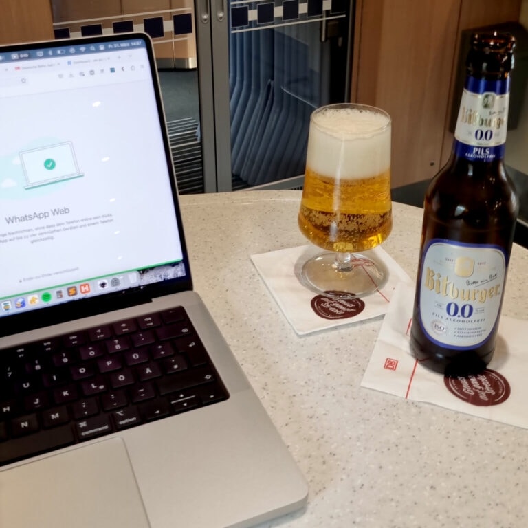A MacBook and a bottle of non-alcoholic beer are standing on a bar table in the on-board bistro of an ICE train. The beer has already been poured into a glass next to it.