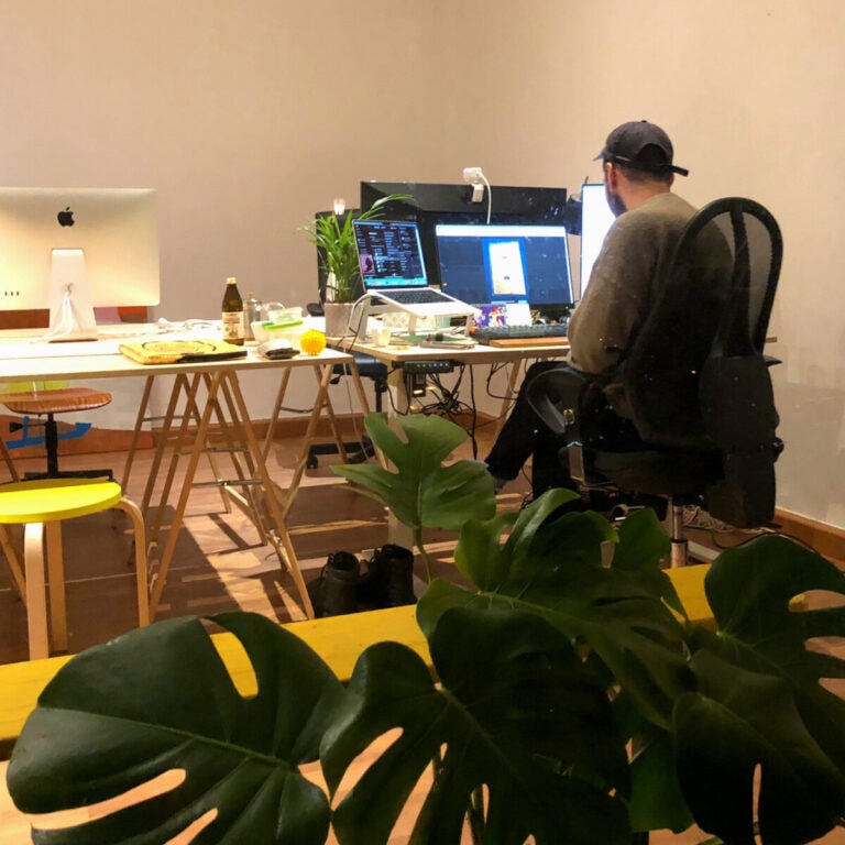 Jeff sits with his back turned to the viewer at a desk with several monitors. In the foreground is a large houseplant.