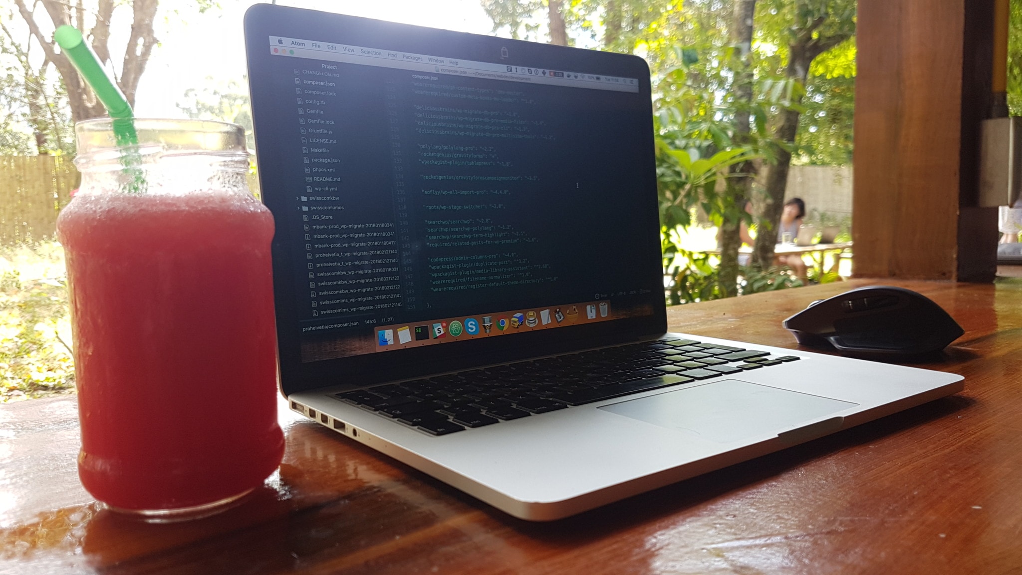 A laptop with a code editor on the screen. Next to it, a refreshing drink and a garden with trees in the background.