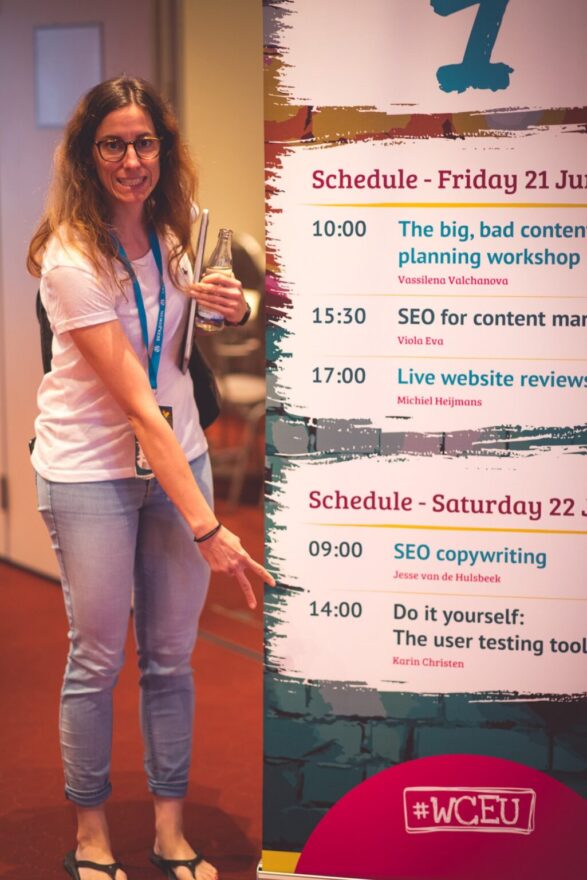 Karin vor einem Konferenz Banner auf welches sie mit dem Finger auf ihren Auftritt zeigt