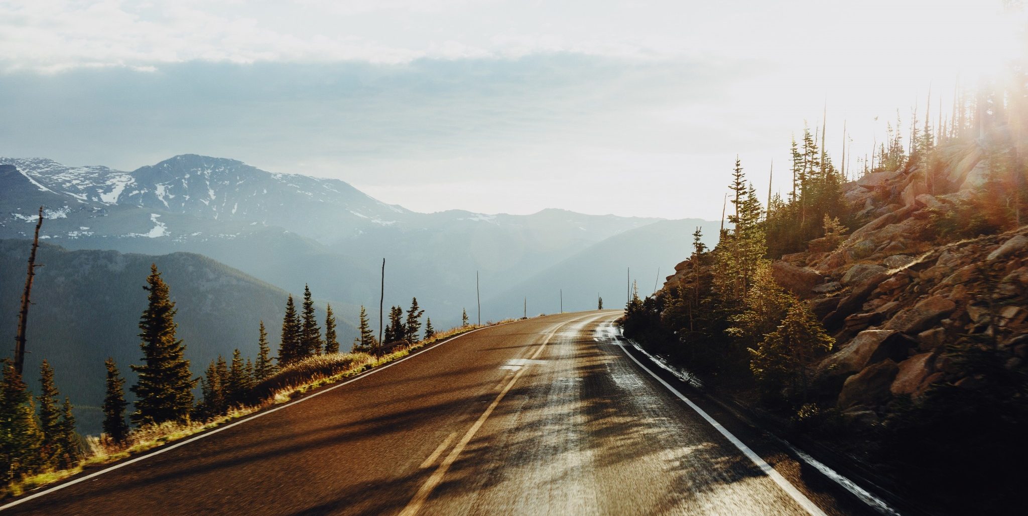 An empty road with mountains in the background. On the left is a cliff, on the right are large boulders and trees