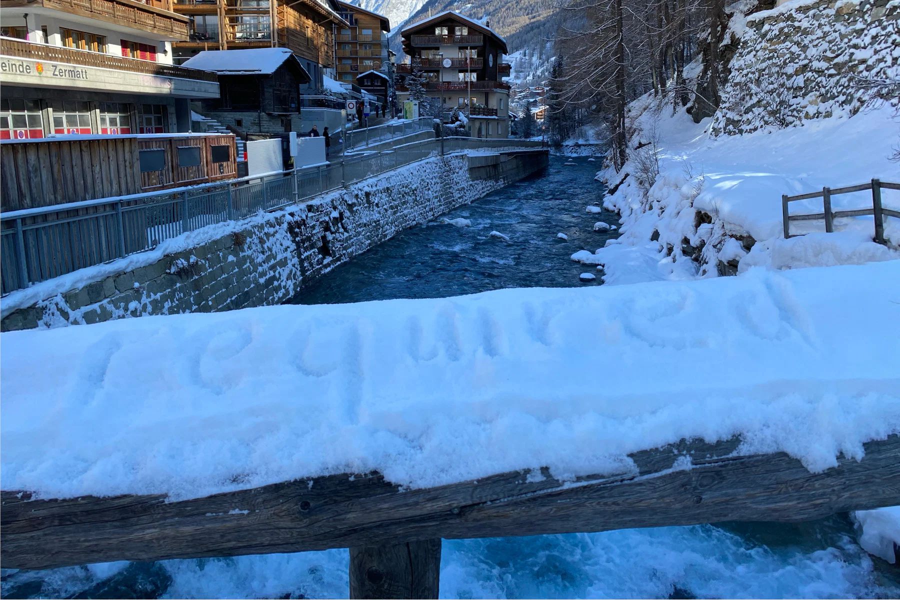 A bridge railing covered in snow, in which the word "require" has been written with a finger. In the background, the village of Zermatt.