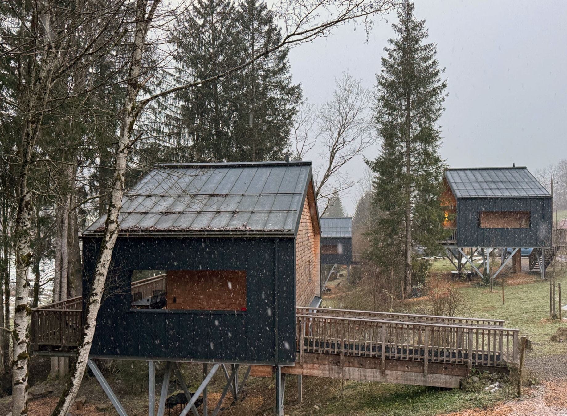Drei Chalets auf Stelzen in winterlicher Landschaft, umgeben von Schnee und Bäumen, mit einer beleuchteten Terrasse, die Gemütlichkeit ausstrahlt.