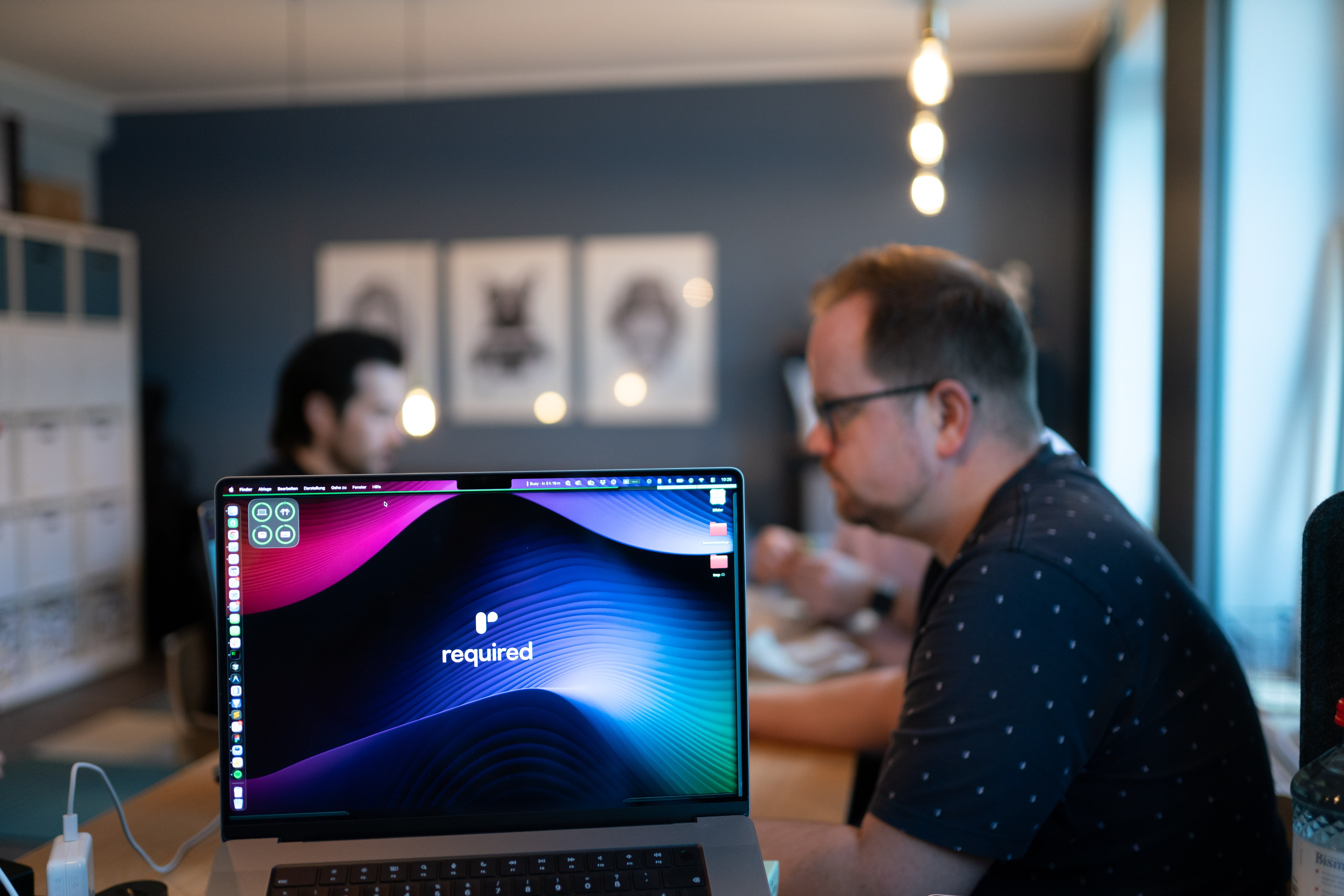 A laptop with the wallpaper of the required.com website. Several employees in the background.