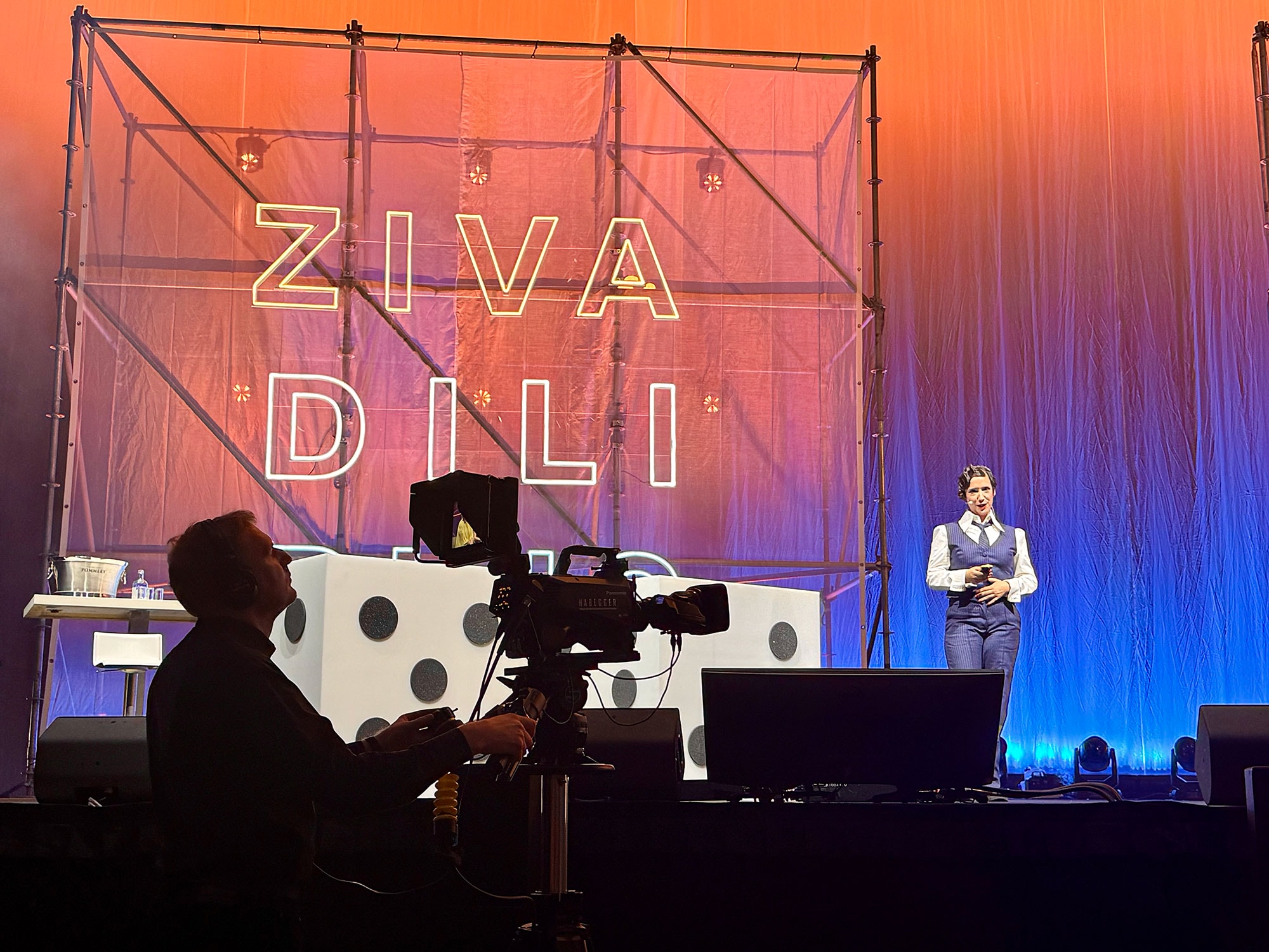Bühnensituation bei einer Veranstaltung: Im Vordergrund filmt eine Kameraperson mit einer professionellen Videokamera. Im Hintergrund steht eine Moderatorin auf der beleuchteten Bühne vor einer grossen Kulisse mit der Aufschrift «ZIVA DILI RING». Die Bühne ist farbig ausgeleuchtet, mit Technik und Requisiten im Veranstaltungssetting.